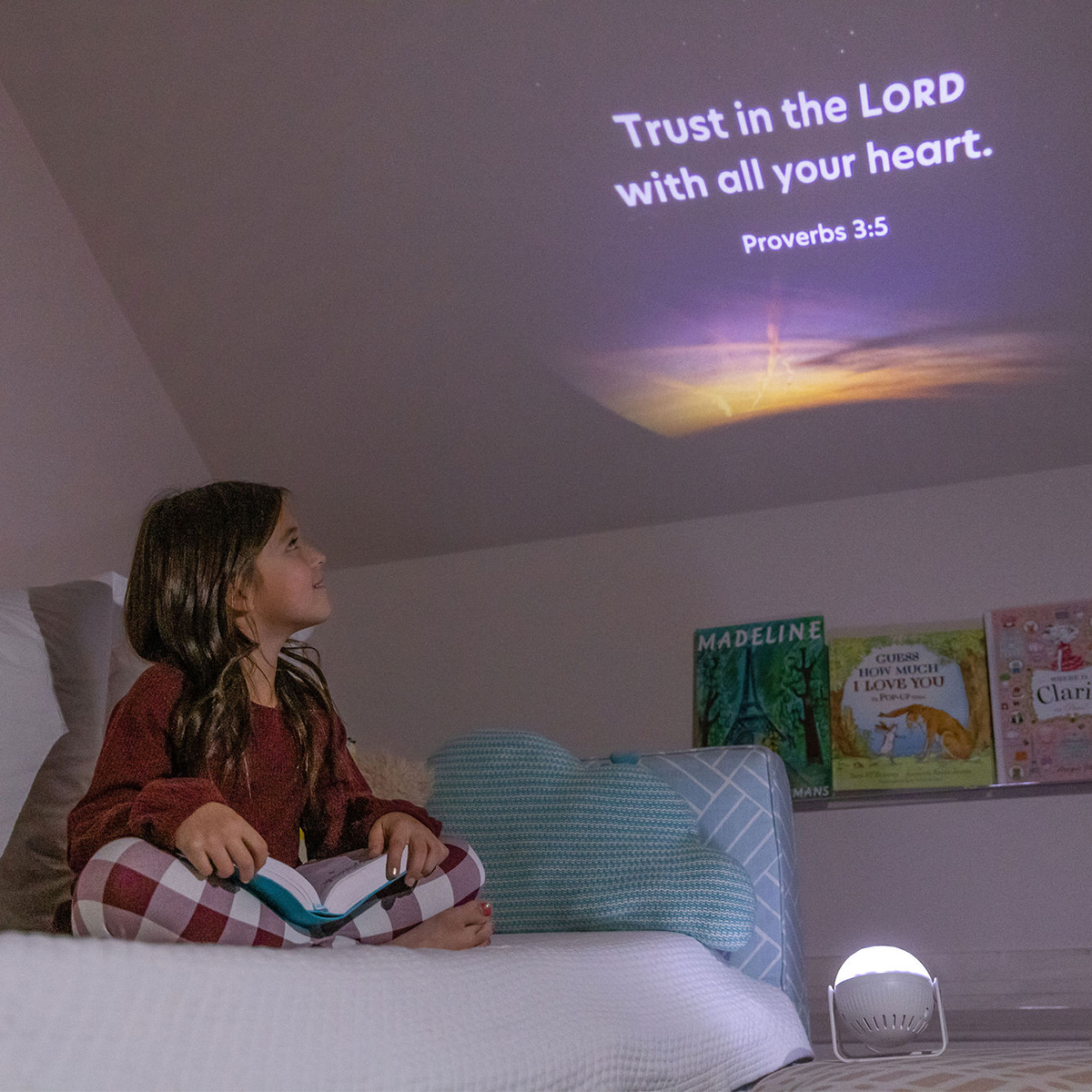 Young 5 year old girl sitting on a bed looking up at the ceiling.  the Glorilight projector is on and shining a night sky image on the ceiling.  The text includes white copy that reads Trust in the Lord with all your heart. Proverbs 3:5.