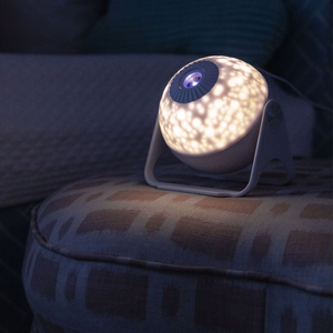 Picture of GloriLight lit up in a dimly lit room. GloriLight sits upon an ottoman with a bed you can barely make out in the background.