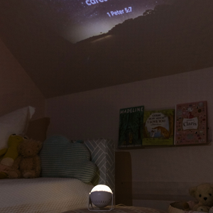 Image of a child's  bedroom with a GloriLight  sitting on an ottoman in the middle of the rooom.  The GloriLight is lit up in the dark rooom and projecting the verse  Cast your cares on Him because He cares for you, first Peter five seven on a slanted ceiling.  Three children's books are on a shelf and stufffed animals are on the bed and in a basket.