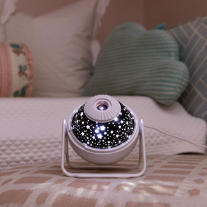 Picture is of a GloriLight with the top cover off. White stars shine from the top of the GloriLight over a black background. The GloriLight appears to be sitting on an ottoman with a bed behind it with four different shaped pillows on top.