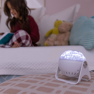 Picture of a GloriLight in the forefront in the bottom right corner. GloriLIight is sitting on an ottoman all lit up. In the background is a young girl reading a book on a bed with pillows and a few stuffed animals.