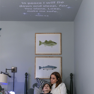 Picture of a kids bedroom with the ceilling lit up with Psalm 4:8, which reads In peace I will lie down and sleep, for You alone, Lord, make me safe. Underneath ceiling is a few framed pics of fish, and underneath the fish there is a  mother and son on a bed. The mom appears to be reading something to her son and the son is looking up at the verse on the ceiling in wonder.  You also can see the top of the GloriLight on the left under a lamp that hangs out of the wall.