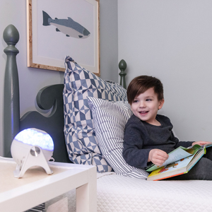 Picture of a four year old boy on his bed with a storybook Bible in his lap. Boy is looking to his right where a GloriLight is lit up on his nightstand. Over top of the bed is a framed picture of a large fish.