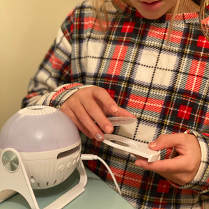 A close-up indoor daytime picture of the GloriLight, the globe nightlight projector. A child in pajamas is loaading a disc into the projector pull out with her hands.  