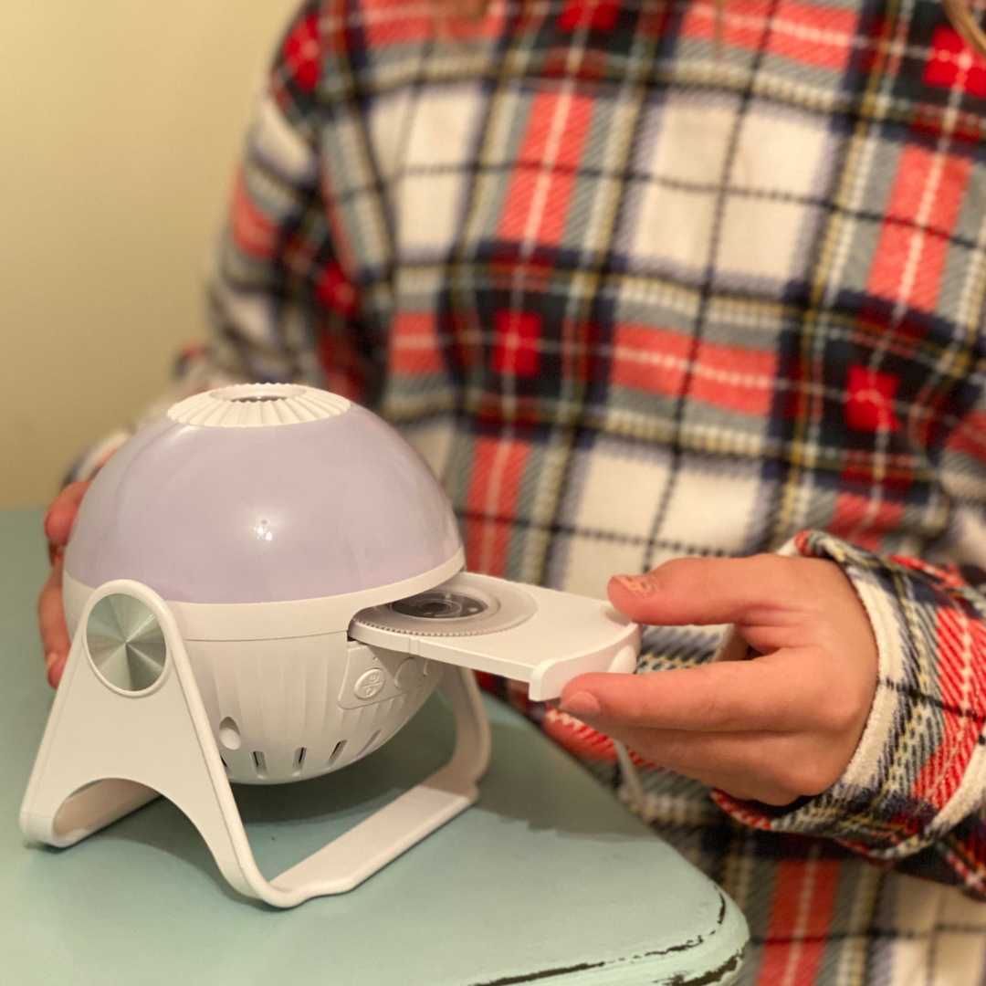 A close-up daytime indoor picture of the GloriLight, the projector globe nightlight. An interchangeable disc is being loaded by a child in pajamas seen from the neck down only. 