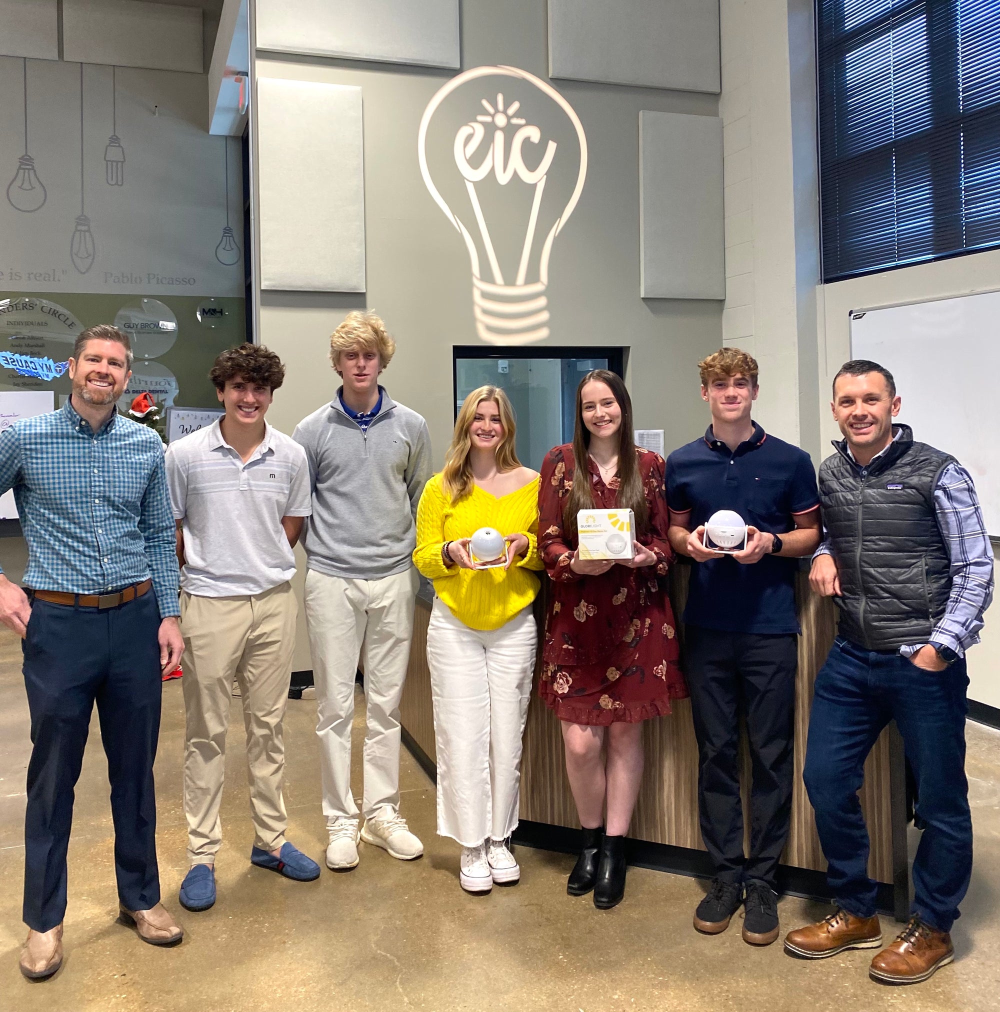 picture of 5 high school students that helped start GlorilLight and co founders Matt Stamper on far left and Clay Banks on far right.  Picture is taken at the EIC with EIC logo in background.  Eic logo is a lightbulb. 