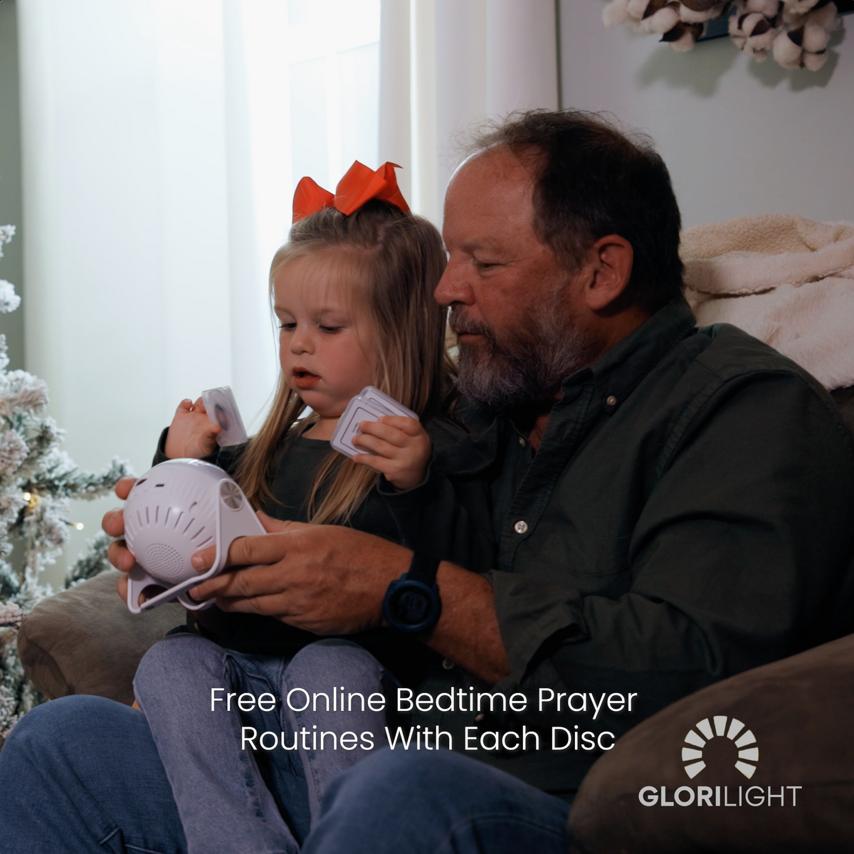 Picture of a grandfather holding his granduaghter on a couch next to a Christmas tree with the GloriLIght and the discs in their hands as they are seemingly figuring how it works. Mid-bottom center text reads, free online bedtime prayer routines with each disc. GloriLight logo and name in the bottom right corner in white.