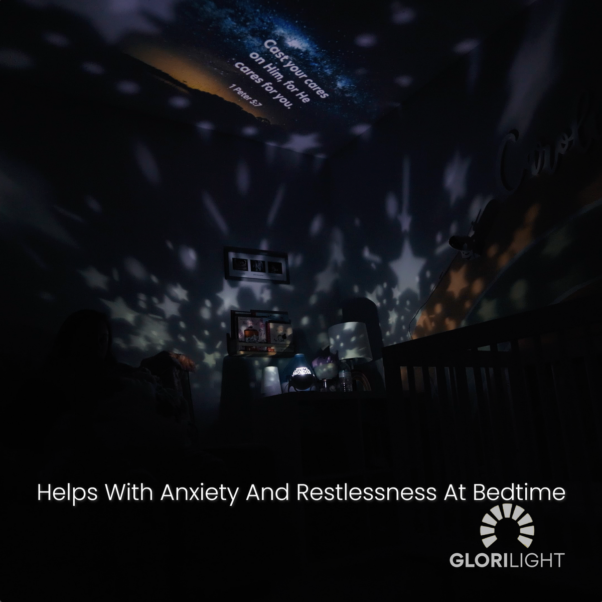 A dark room shot with the GloriLight projection lighting up the ceiling with 1 Peter 5:7 on it. Stars are also being projected around the room and text at the bottom center reads, helps with anixiety and restlessnes at bedtime. The GloriLight logo and name are bottom right in white. 