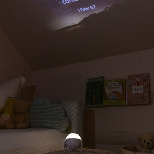 Image of a child's  bedroom with a GloriLight  sitting on an ottoman in the middle of the rooom.  The GloriLight is lit up in the dark rooom and projecting the verse  Cast your cares on Him because He cares for you, first Peter five seven on a slanted ceiling.  Three children's books are on a shelf and stufffed animals are on the bed and in a basket. 
